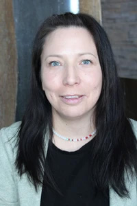 The image shows a woman with dark hair wearing a necklace, smiling at the camera. She has light skin and appears to be indoors, possibly in an office setting.
