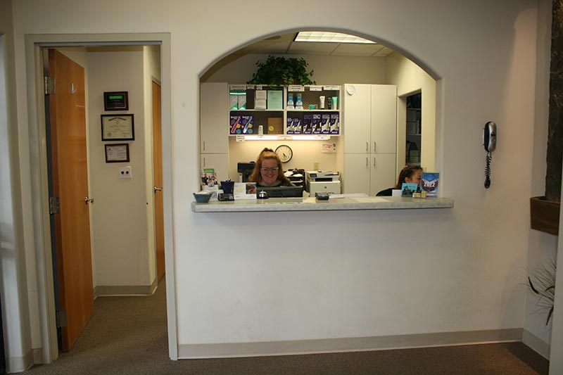 The image shows an interior view of a reception area with a woman standing behind a counter, which has a sign on top indicating  Check-In  and various items such as books and cards on display. A wall clock is visible above the counter, and there are shelves with bottles and other items to the right. The room appears to be well-lit and clean.