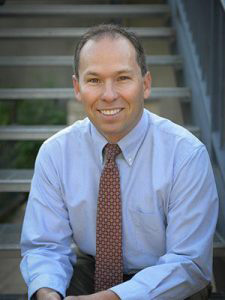 A man wearing a blue shirt and tie, sitting on stairs with his hands on his knees.