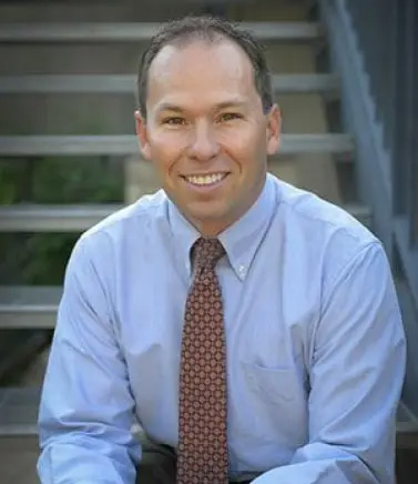 The image shows a man sitting on steps with a smile, wearing a blue shirt and a tie, posing for a portrait-style photograph.