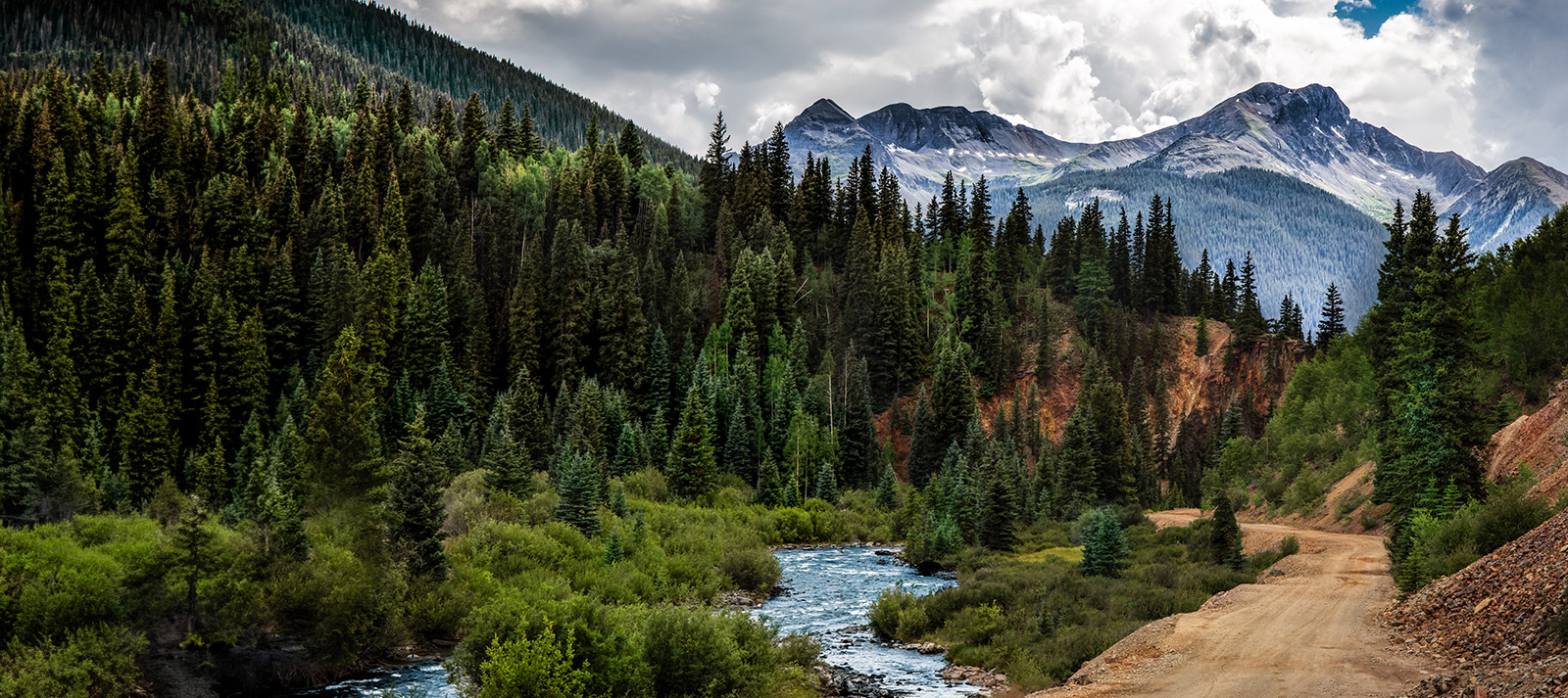 The image shows a scenic mountain landscape with a winding road through a forested area, featuring tall evergreen trees, a river, mountains in the background, and a clear sky above.