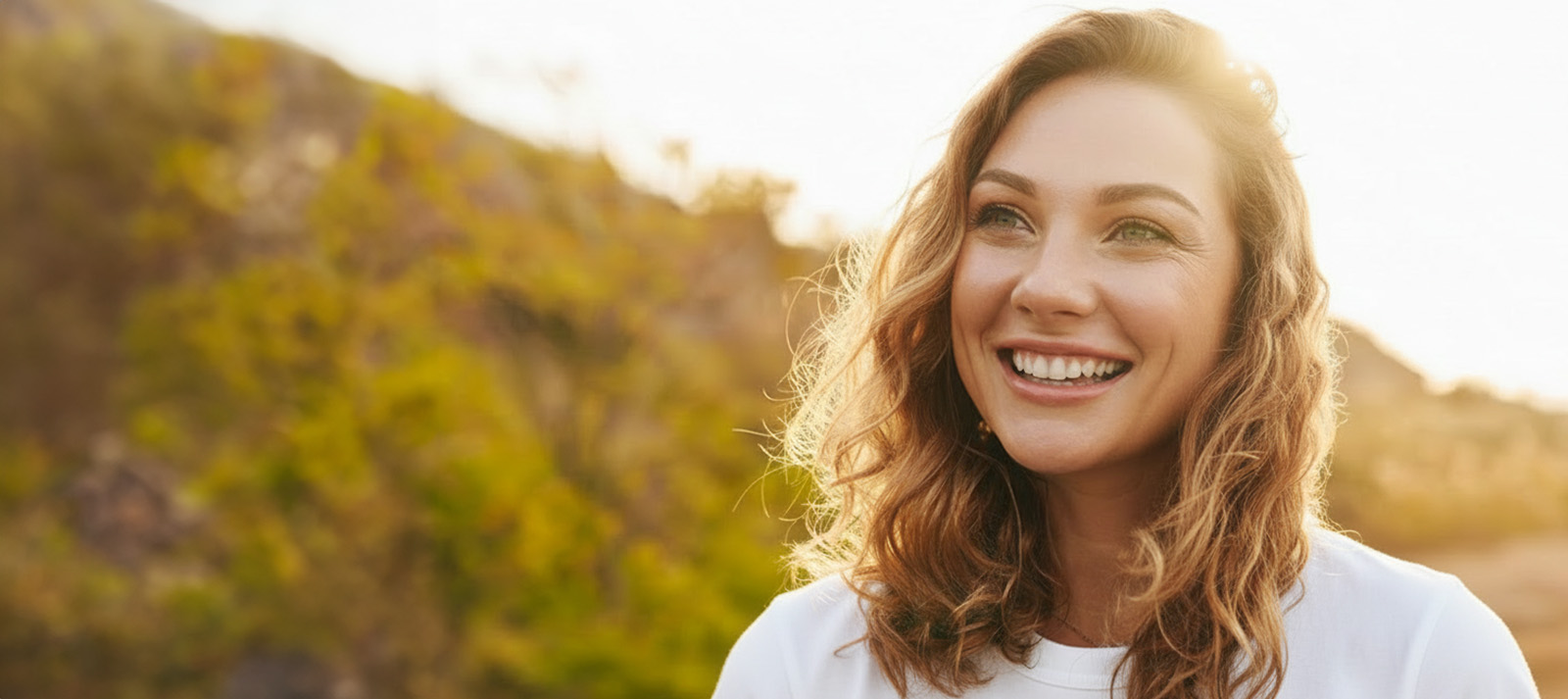 A young woman with curly hair smiles at the camera while standing outdoors during sunset.