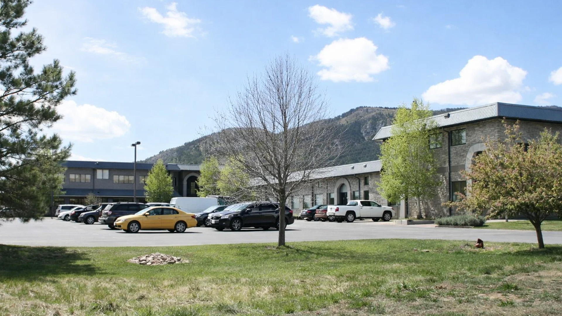 The image shows a parking lot with several cars parked in front of a building during daytime under clear skies.