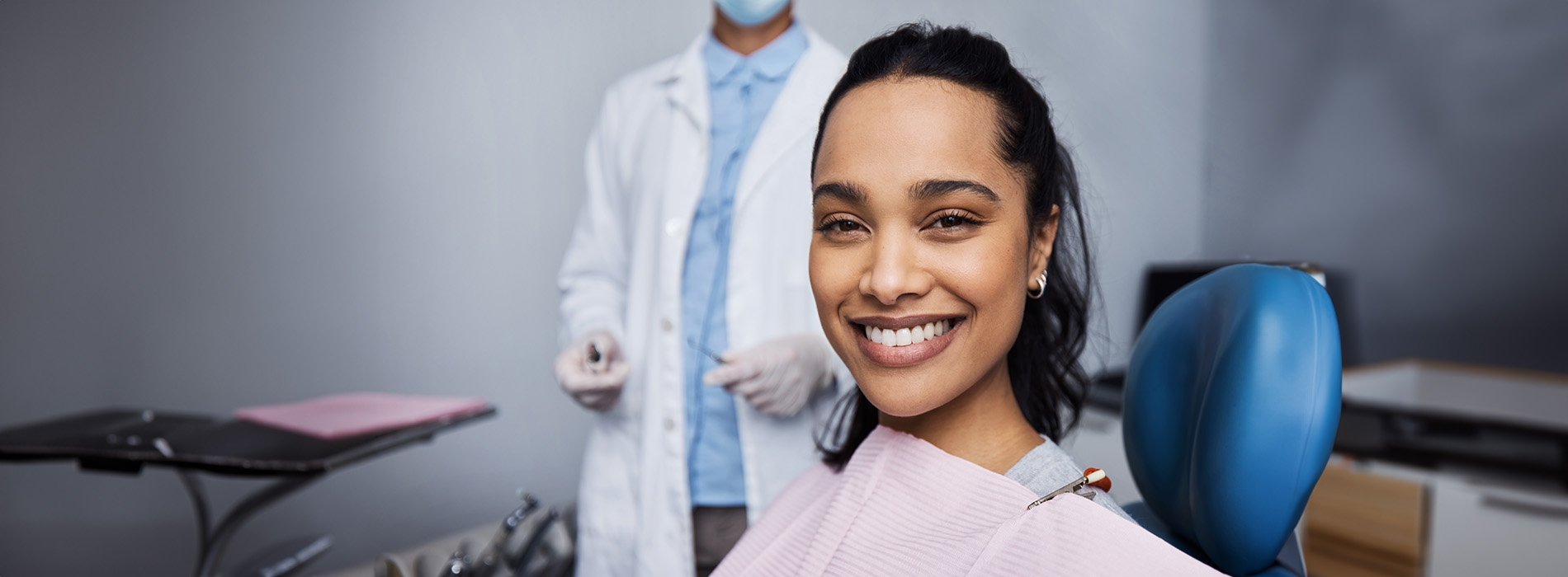 The image shows a person smiling at the camera while seated in a dental chair, with a dental professional standing behind them holding a dental mirror, both wearing surgical masks and in a dental office setting.