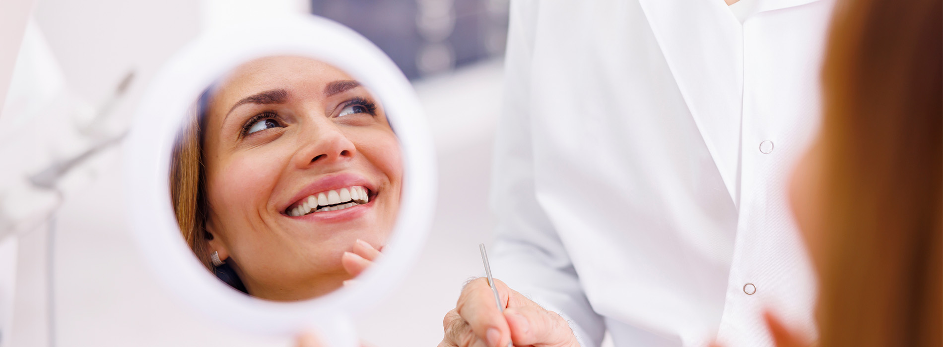 The image shows a woman smiling at her reflection in a mirror while being attended to by a professional with dental tools, likely in a dental office setting.