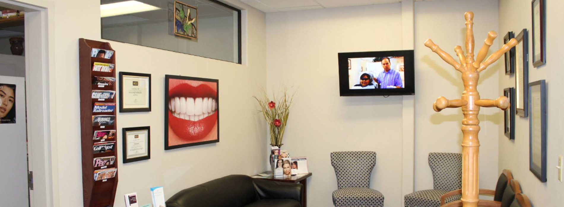 The image shows an interior view of a dental office reception area with a modern design featuring a comfortable seating arrangement, a television displaying a news program, and framed artwork on the walls.
