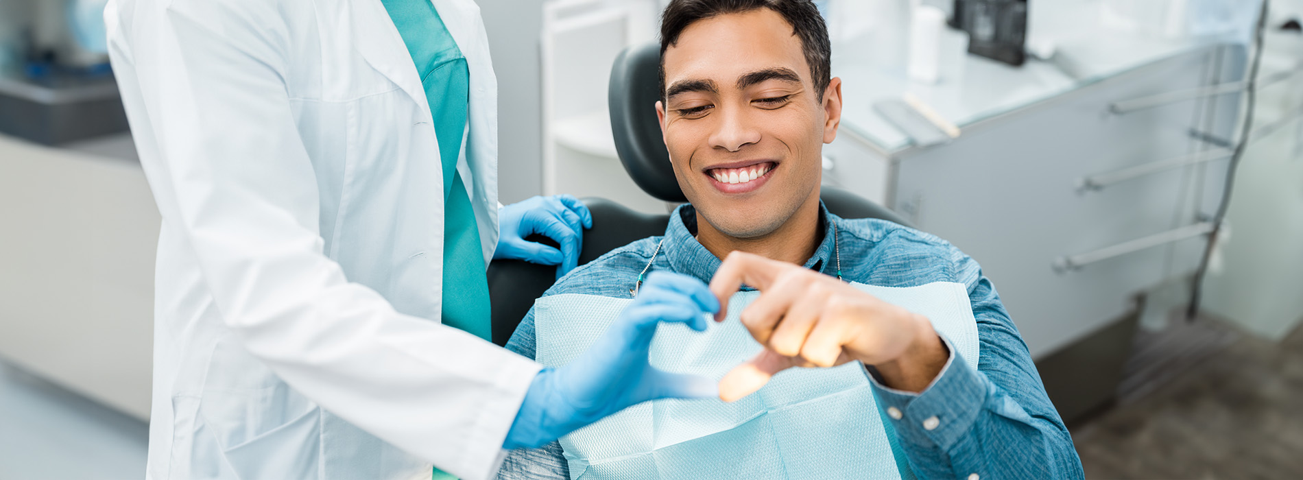 A man wearing a blue surgical mask is seated in a dental chair with a smile on his face while being attended to by a dental professional, who appears to be in mid-action of cleaning teeth.