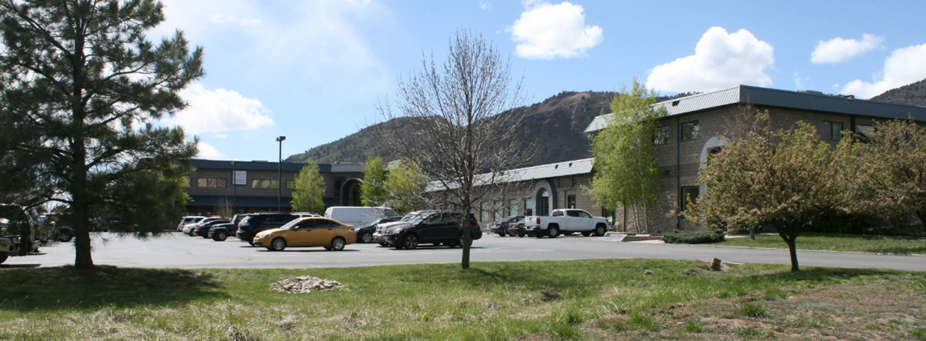 The image shows a panoramic view of a parking lot with multiple cars parked alongside a building, under a clear blue sky, with trees visible in the background.