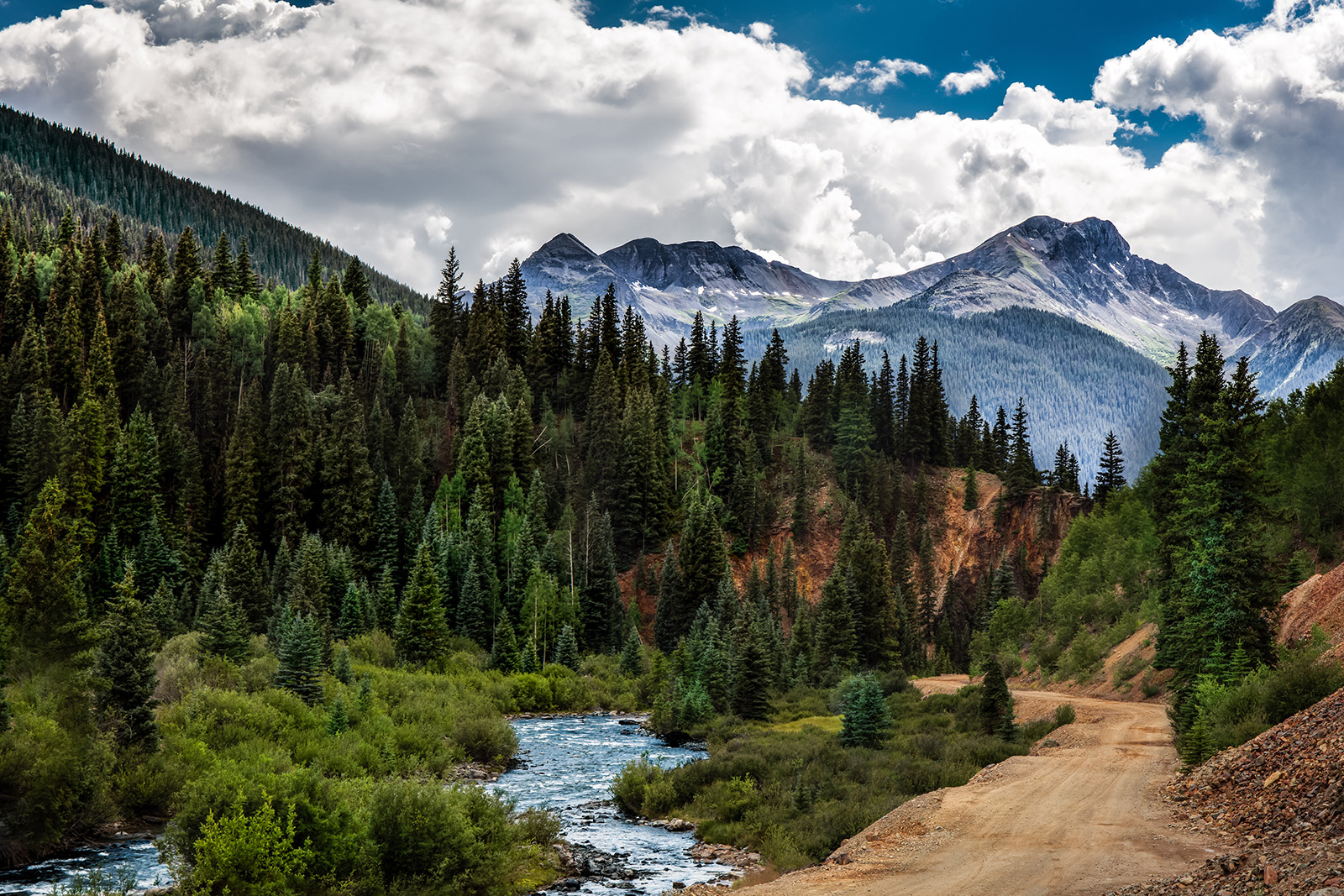 The image shows a scenic mountain landscape with a forested area, a winding dirt road, and mountains in the background under a partly cloudy sky.