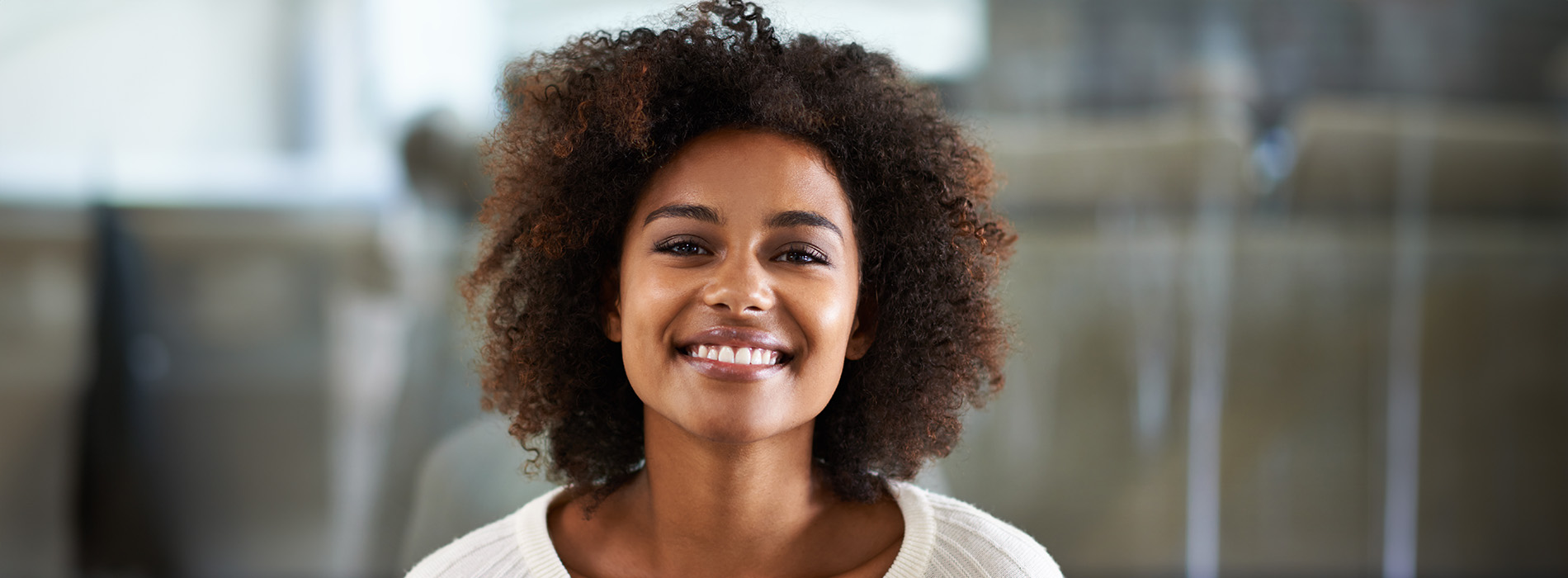 A young woman with curly hair smiles at the camera, wearing a white top against a blurred background.