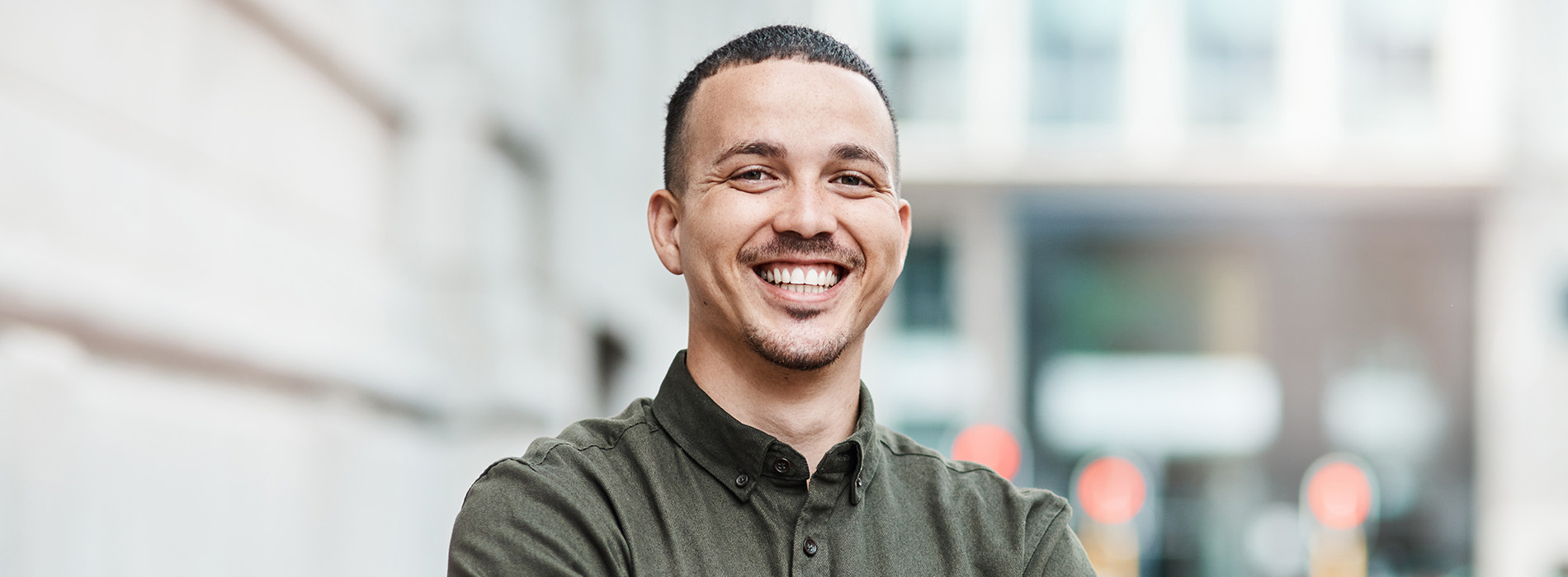 The image shows a man standing with his arms crossed, smiling at the camera, against an urban background.