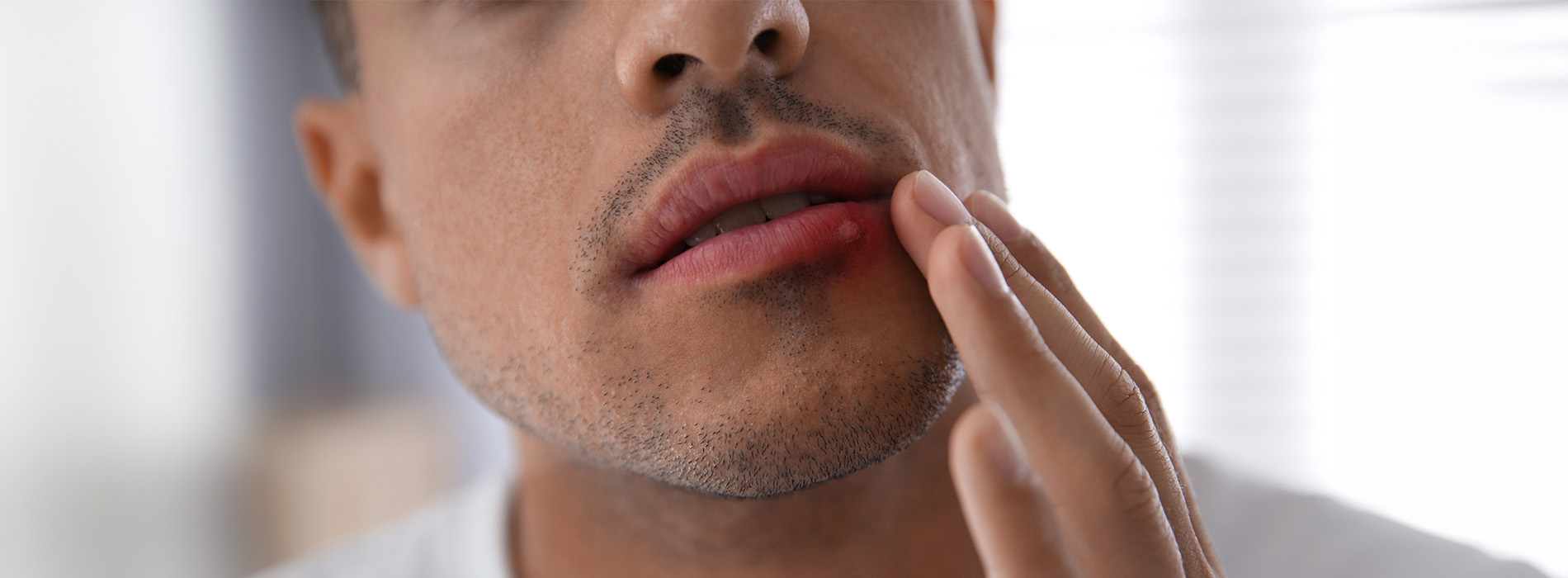 The image shows a close-up view of a person applying a substance to their face with a visible reflection on a surface behind them, set against a blurred background.