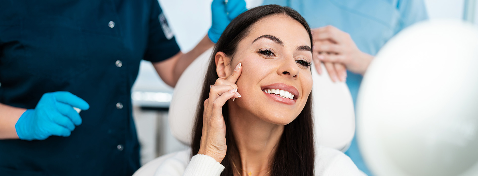A woman with her eyes closed, smiling broadly, seated in a dental chair during a dental procedure, surrounded by dental professionals wearing masks and gloves.
