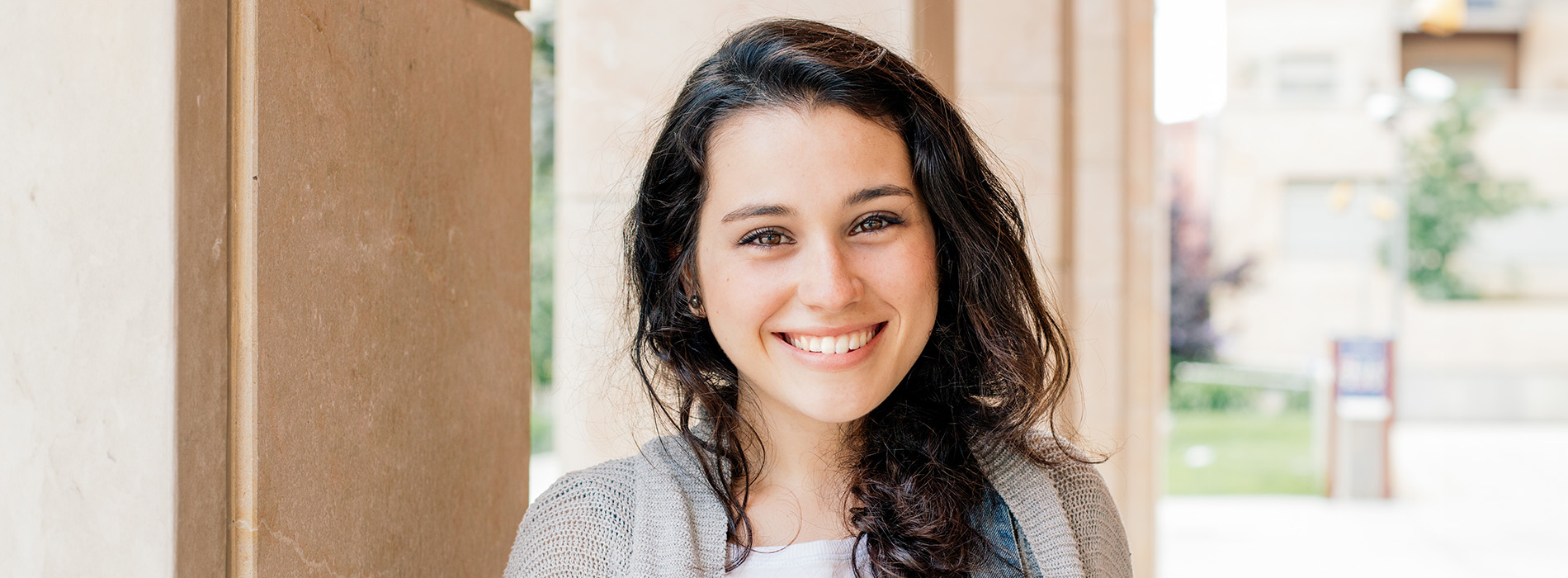 The image features a woman with a smile, standing outdoors with a building behind her, wearing a casual top and holding a small object in her hand.
