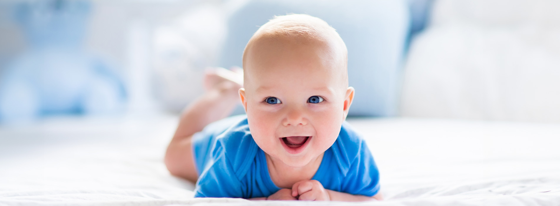 A smiling baby lying on its back with hands near face, wearing blue clothing, set against blurred background.