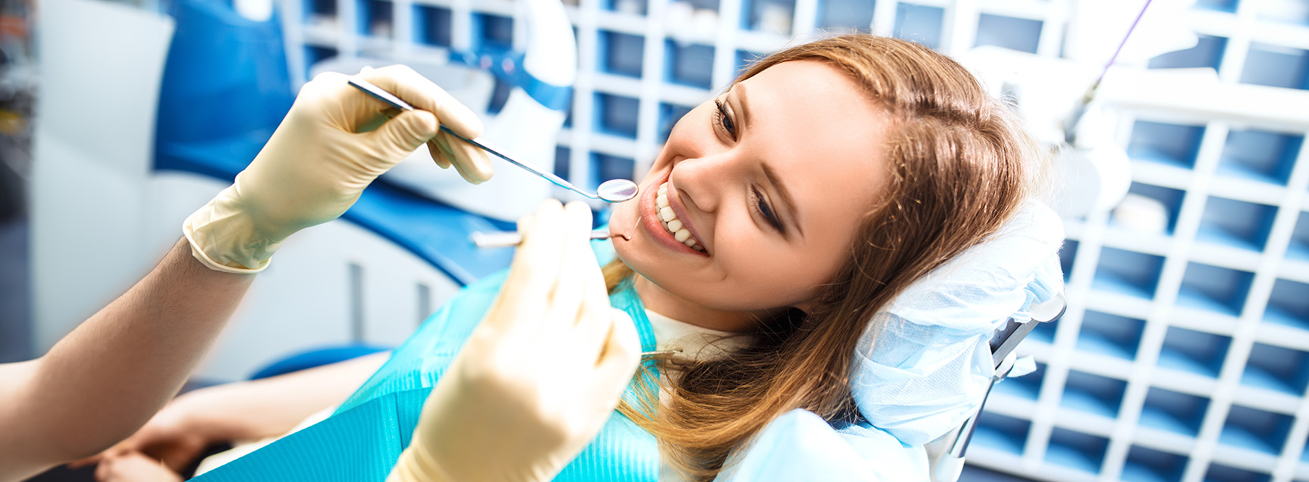 A woman sitting in a dental chair with a smile, wearing a blue surgical gown and holding a toothbrush, being attended to by a dentist who is cleaning her teeth while looking at her with a smile.