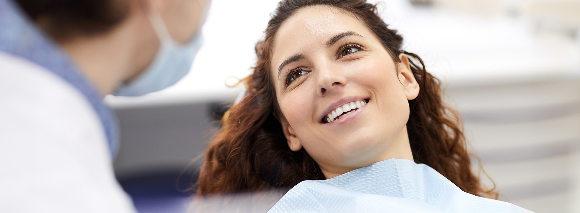 The image shows a woman with a radiant smile sitting in a dental chair, receiving dental care from a professional wearing white protective gear, with both of them appearing happy and engaged in their respective roles.