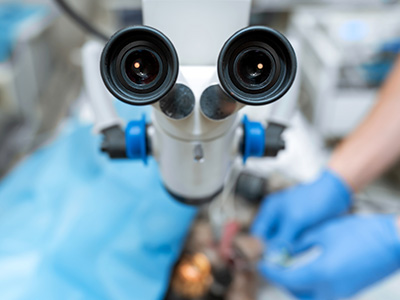 The image shows a person working with medical equipment, possibly in an operating room or medical facility, examining a microscope with a camera attached to it, focusing on something that appears to be under examination.