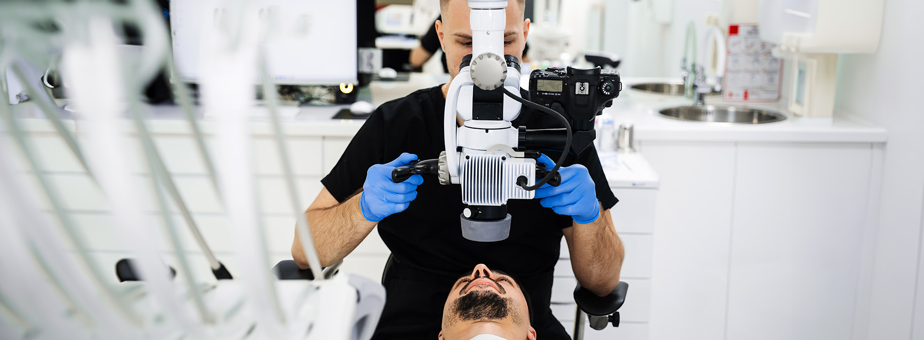 The image shows a man with dark hair wearing a black shirt and blue gloves holding a camera, taking a photograph of his reflection in a mirror while standing in front of a sink.