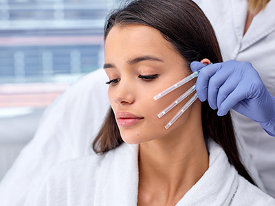 The image shows a woman with long hair receiving a facial treatment from a professional using a device that appears to be for skin care, possibly involving a machine or tool, while wearing a white lab coat.