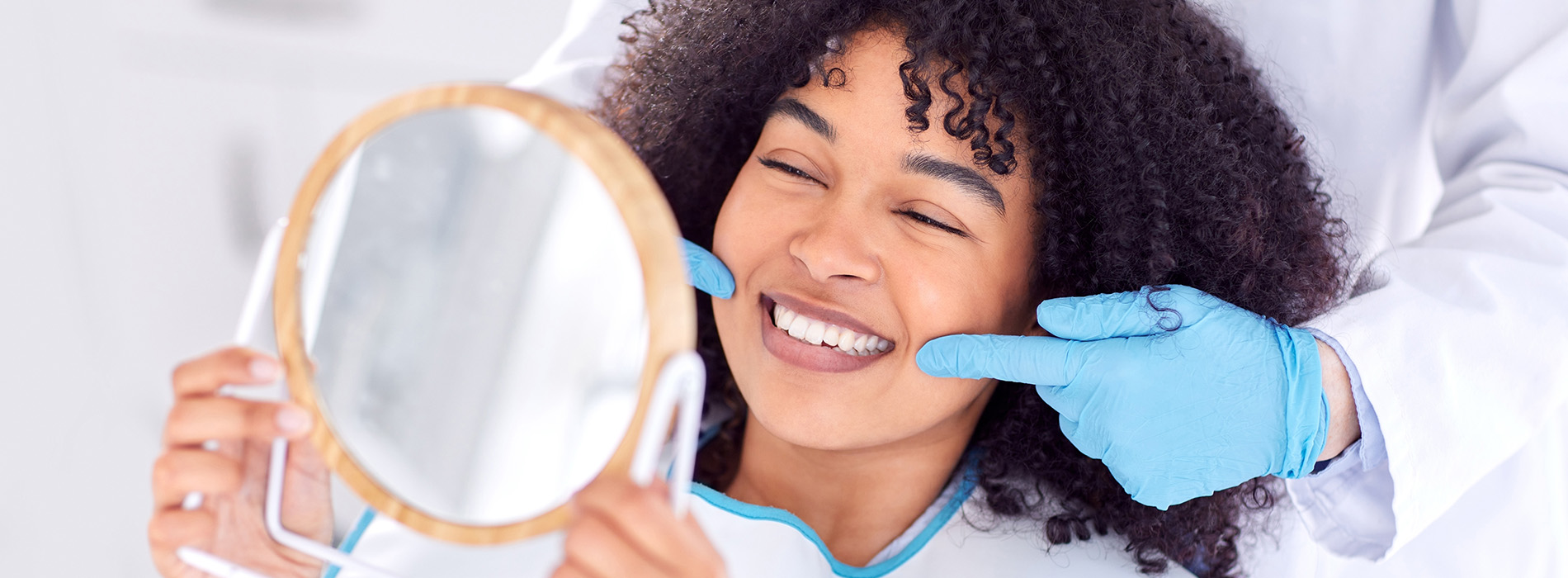 A smiling woman with curly hair holding a magnifying glass over her face while looking into a mirror.