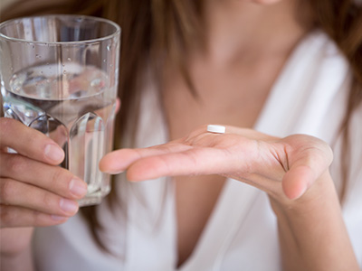 The image features a woman holding a pill between her fingers, with two glasses of water placed on either side of the pill, against a blurred background.