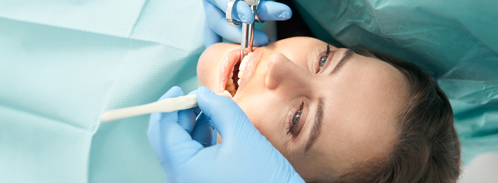 The image shows a person receiving dental treatment with a dentist using tools and wearing gloves, set against a backdrop of medical equipment and supplies.