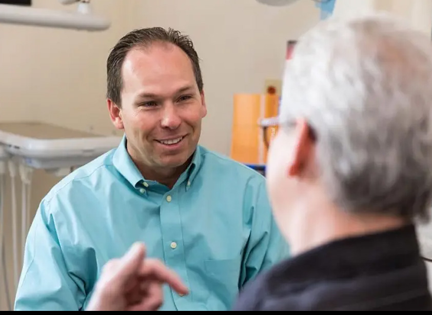 A man wearing a blue shirt is sitting in front of another man who is standing behind him, both are indoors with medical equipment visible in the background.