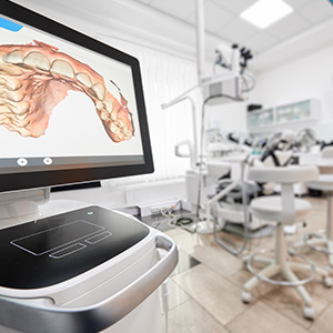 The image shows a dental office setting with a large screen displaying an X-ray of a human mouth and teeth, along with a 3D model or rendering of the same mouth on a computer monitor. There is also a dental chair visible in the background.