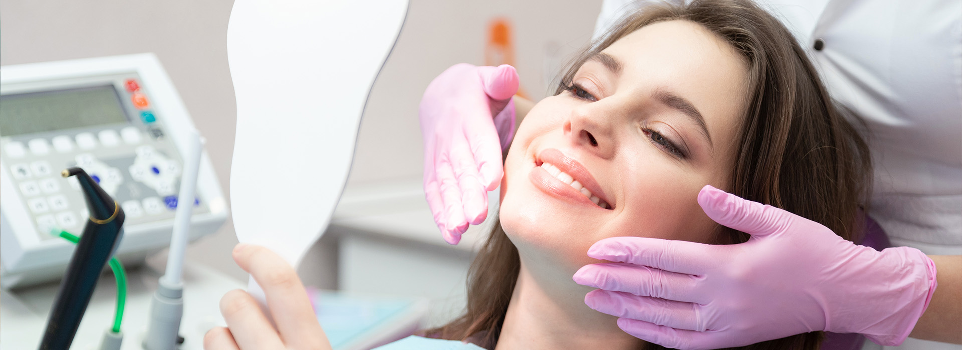 The image depicts a woman sitting in a dental chair with a hygienist performing an oral examination, using a mirror to inspect her teeth while she smiles.
