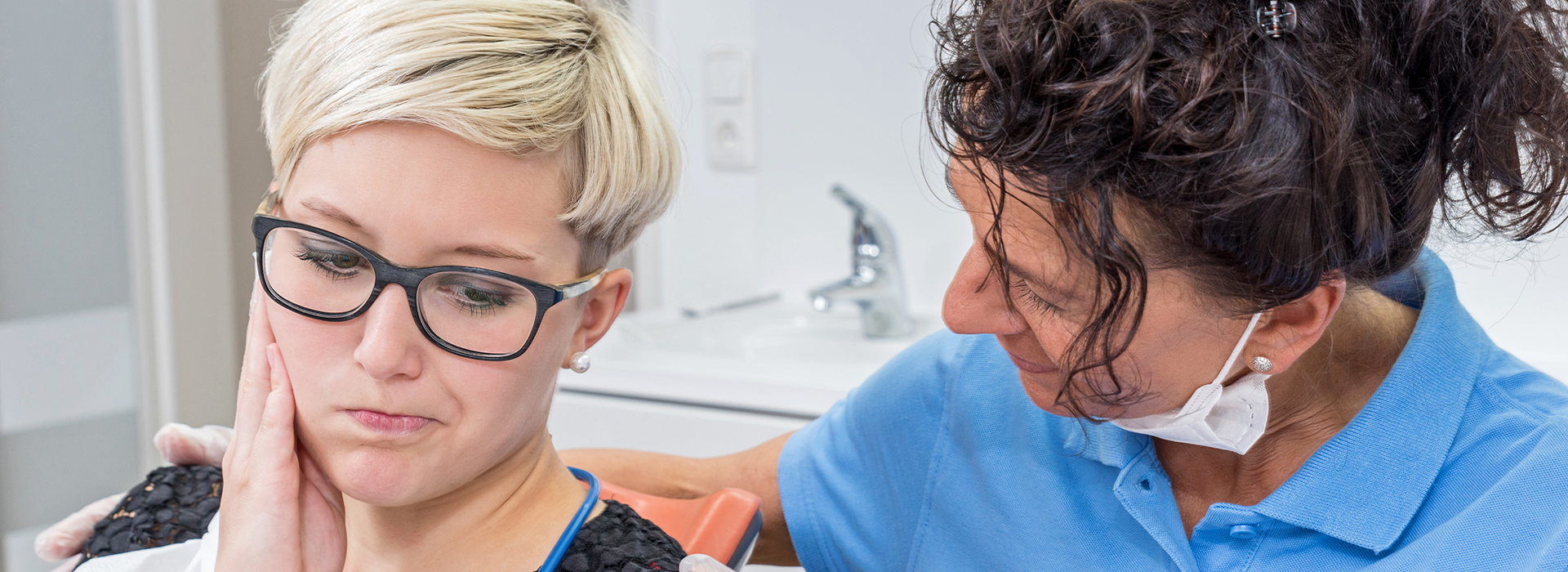 An image of a dental care professional assisting a patient with an oral examination, featuring a woman wearing glasses and another individual with a stethoscope.