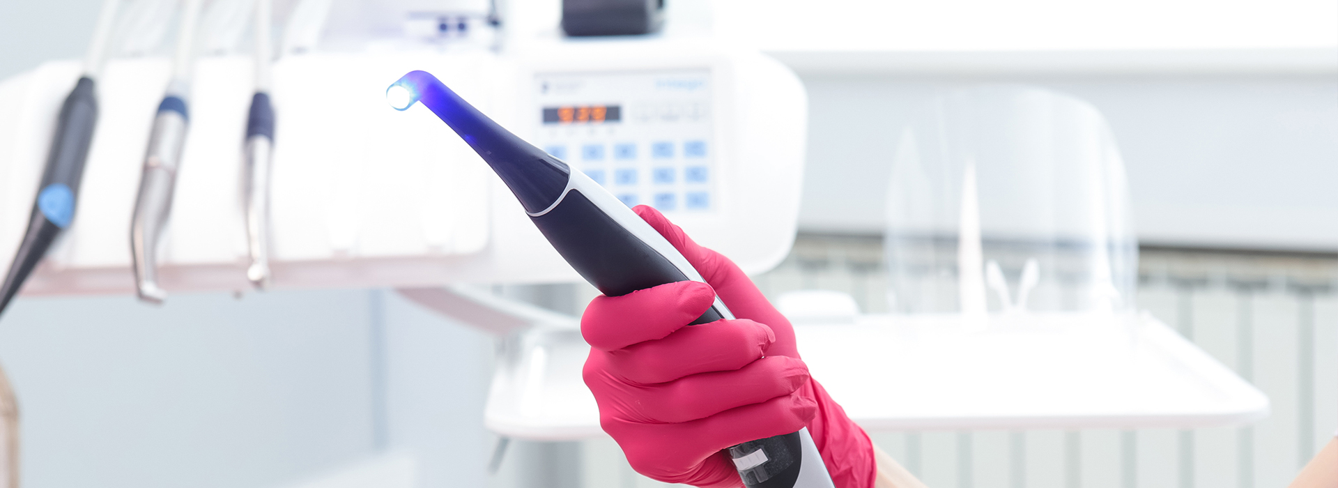 The image shows a close-up of a person s hand holding an electronic device with a blue light, wearing pink gloves, and standing in front of a backdrop that includes various pieces of laboratory equipment.