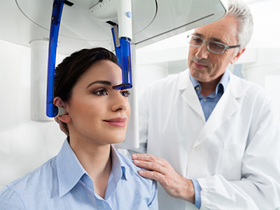 In the image, a woman is seated with her head inside a medical device while a man in a white lab coat stands beside her, observing the equipment.