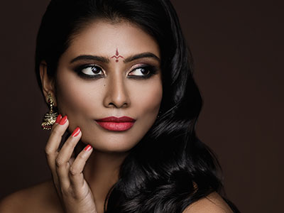 The image features a woman with dark hair and makeup, wearing traditional attire and jewelry, posing for a portrait with her hand on her chin against a neutral background.