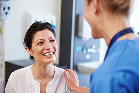 A woman in a white coat smiles at another woman who seems surprised or excited while they are both in a medical setting, with a doctor standing between them.