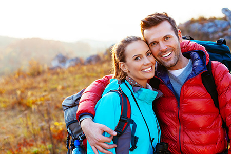 The image shows a man and woman standing close together outdoors during daytime, with the man wearing a backpack and both smiling at the camera.