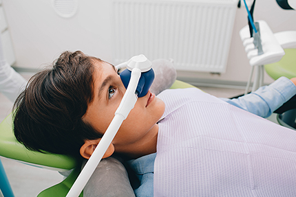A young person wearing a medical mask and sitting in a dental chair with a dental mirror on their face.