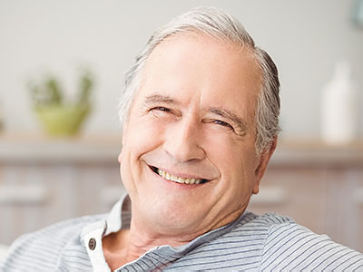 The image shows a smiling elderly man with white hair, wearing a blue shirt, sitting comfortably with his arm resting on a surface.