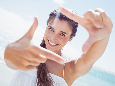 The image shows a smiling woman taking a selfie with her hand, standing on a beach with clear skies and bright sunlight.